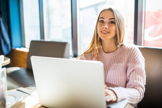 Hübsches blondes Mädchen im weißen Pullover der Mode bei der Arbeit an ihrem Laptop im Café in der täglichen Zeit