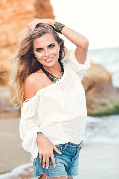 Hübsches blondes Mädchen mit langen Haaren posiert zur Kamera am Strand nahe Meer.