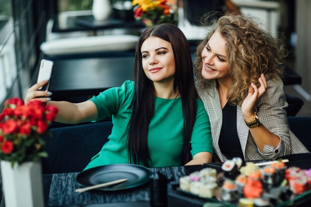 Hübsches blondes und brünettes Mädchen, das ein Foto am Handy mit Sushi-Teller auf dem Tisch macht. Chenese essen, Freunde Zeit.