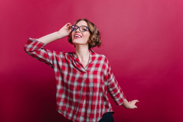 Hübsches brünettes Mädchen im Freizeithemd, das positive Emotionen auf Rotweinwand ausdrückt. fröhliche lockige junge Dame in Gläsern.