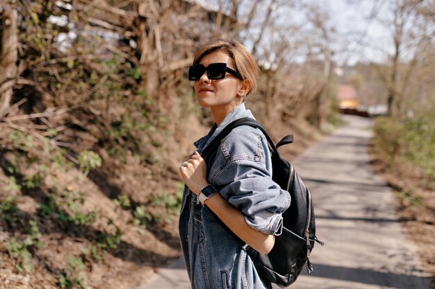 Hübsches charmantes Mädchen mit hellem Haar in Sonnenbrille und Jeansjacke mit Rucksack geht an warmen Frühlingstagen auf dem Waldweg spazieren