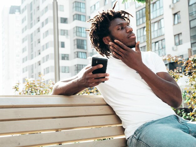 Hübsches Hipster-ModelUnrasierter afrikanischer Mann in weißem Sommer-T-ShirtModemann mit Dreadlocks-Frisur Posiert auf der StraßeMit Handy-Apps auf Smartphone-Bildschirm schauen