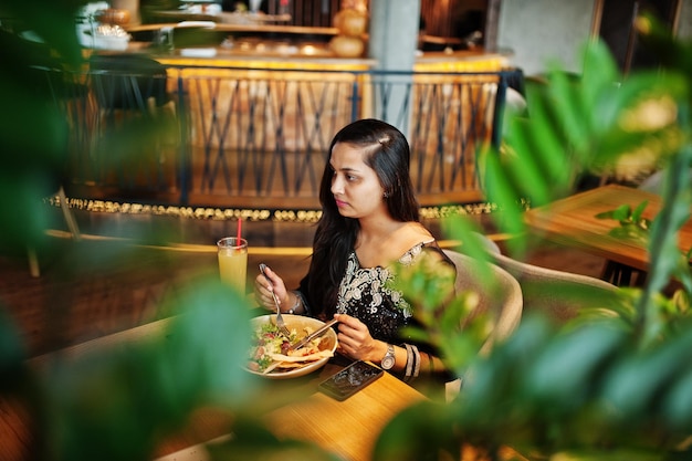 Hübsches indisches Mädchen in schwarzem Saree-Kleid posierte im Restaurant, das mit Saft und Salat am Tisch saß