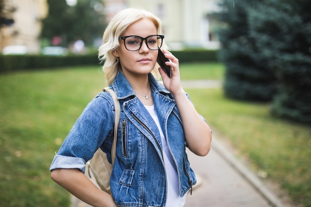 Hübsches junges blondes Mädchen benutzt Telefon, um soziale Netzwerkkonversation im Herbstmorgen der Stadt zu scrollen
