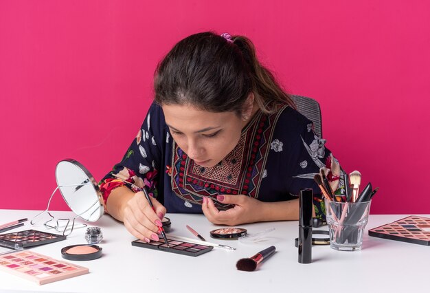 Hübsches junges brünettes Mädchen, das am Tisch mit Make-up-Tools sitzt und Eyeliner hält und die Lidschattenpalette betrachtet