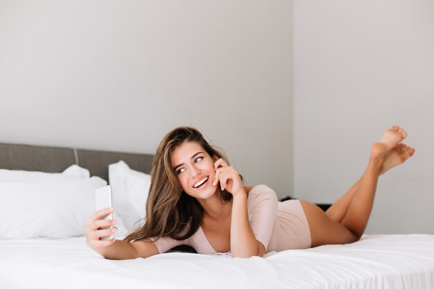 Hübsches junges Mädchen im Pyjama, das morgens auf Bett in Wohnung liegt. Sie macht ein Selfie-Porträt am Telefon und sieht begeistert aus.