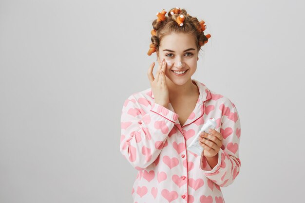 Hübsches junges Mädchen in Lockenwicklern und Pyjama tragen Gesichtscreme auf