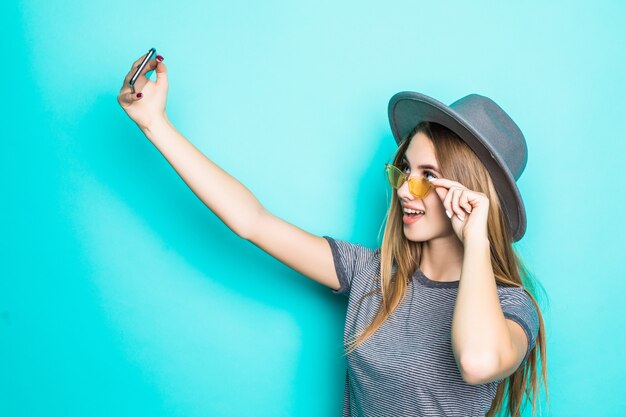 Hübsches junges Modell im Mode-T-Shirt, im Hut und in der transparenten Brille mit Telefon in ihren Händen macht Selfie lokalisiert auf grünem Hintergrund