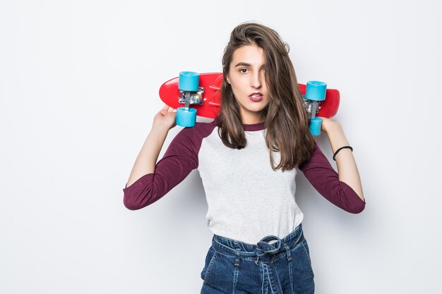 Hübsches junges Skatermädchen, das rotes Skateboard auf ihrem Rücken lokalisiert auf weißer Wand hält