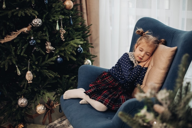 Hübsches kaukasisches Kind in einem Kleid schläft nahe dem Weihnachtsbaum auf einem blauen Sessel zu Hause