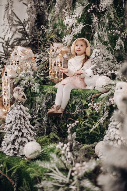 Hübsches kaukasisches Kind mit langen blonden Haaren lächelt und sitzt in der Weihnachtsatmosphäre mit vielen geschmückten Bäumen um sie herum und einem kleinen Kaninchen