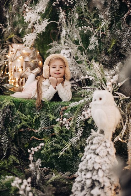 Hübsches kaukasisches Kind mit langen blonden Haaren liegt in der Weihnachtsatmosphäre mit vielen geschmückten Bäumen um sie und Eule