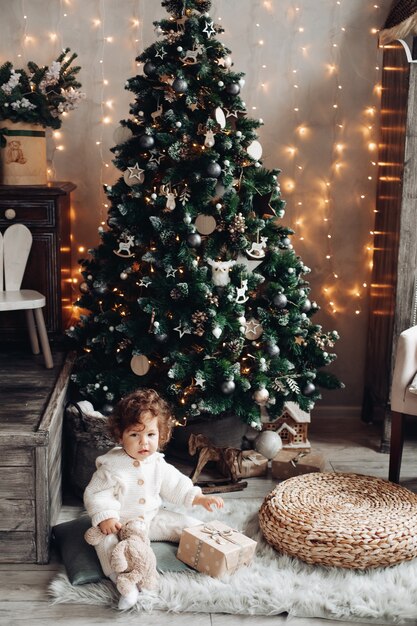 Hübsches kaukasisches Mädchen mit einem lockigen Haar sitzt nahe einem großen schönen Weihnachtsbaum