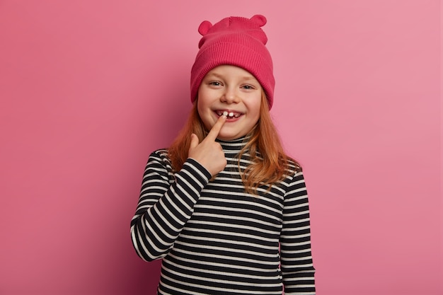Hübsches kleines Ingwermädchen zeigt ihrem ersten Erwachsenen zwei Zähne, lacht und freut sich, drückt positive Gefühle aus, hält den Mund offen, bereitet sich auf die mündliche Untersuchung vor, gekleidet in einen gestreiften Pullover und einen rosa Hut