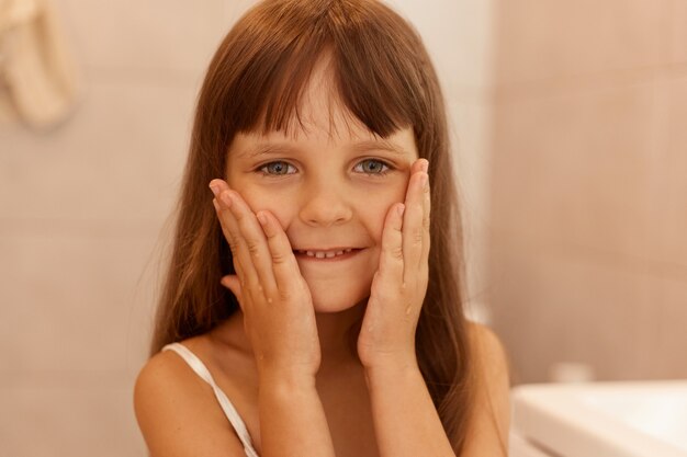Hübsches kleines Mädchen, das Wangen berührt, die Kamera mit einem angenehmen Lächeln und positiven Emotionen betrachtet, nach Hygiene- und Schönheitsverfahren im Badezimmer posiert.