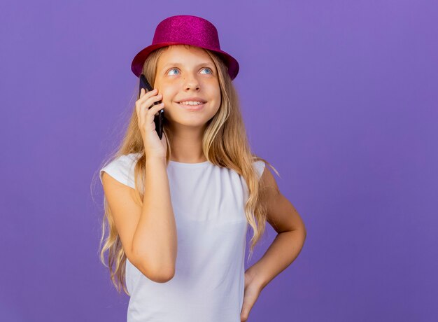 Hübsches kleines Mädchen im Feiertagshut, der auf lächelndem Handy spricht, Geburtstagsfeierkonzept, das über lila Hintergrund steht