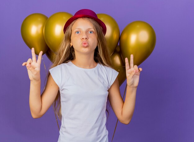 Hübsches kleines Mädchen im Feiertagshut mit Bündel der Luftballons, die Kamera glücklich und positiv betrachten, die V-Zeichen, Geburtstagsfeierkonzept über lila Hintergrund stehend zeigt