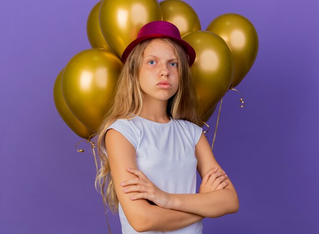 Hübsches kleines Mädchen im Feiertagshut mit Bündel von Ballons, die Kamera mit ernstem Gesicht betrachten, das unzufrieden ist, Geburtstagsfeierkonzept, das über lila Hintergrund steht