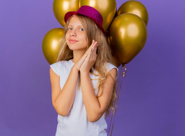 Hübsches kleines Mädchen im Feiertagshut mit Bündel von Ballons, die Palmen zusammenhalten, die auf Überraschung warten, Geburtstagsfeierkonzept stehen über lila Hintergrund