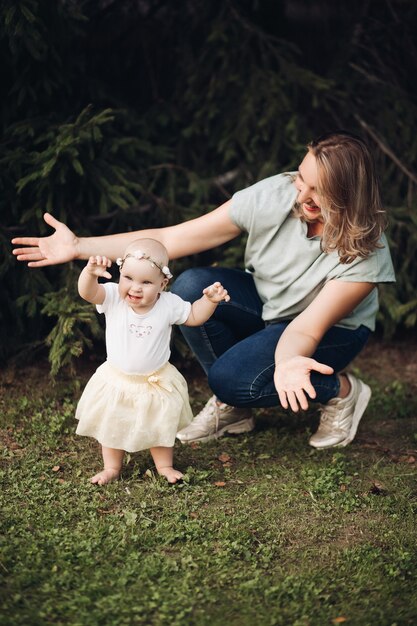 Hübsches kleines Mädchen mit kurzen blonden Haaren und hübschem Lächeln im weißen Kleid sitzt im Sommer mit ihrer Mutter auf einem Gras im Park