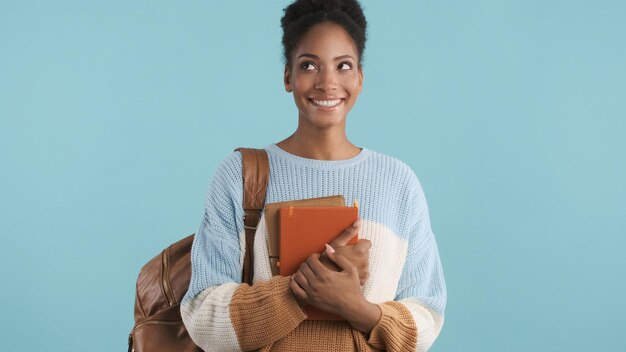 Hübsches lächelndes afrikanisch-amerikanisches Studentenmädchen mit Büchern, die freudig über farbenfrohen Hintergrund beiseite schauen