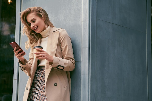 Hübsches lächelndes blondes Mädchen im stylischen Trenchcoat mit Kaffee zum Mitnehmen freudig mit dem Handy im Freien