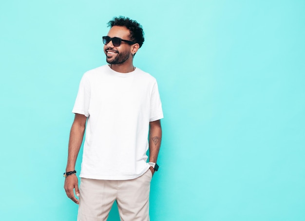 Kostenloses Foto hübsches lächelndes hipster-model sexy unrasierter mann in weißem sommert-shirt und jeanskleidung modemann posiert in der nähe der blauen wand im studio isoliert mit sonnenbrille