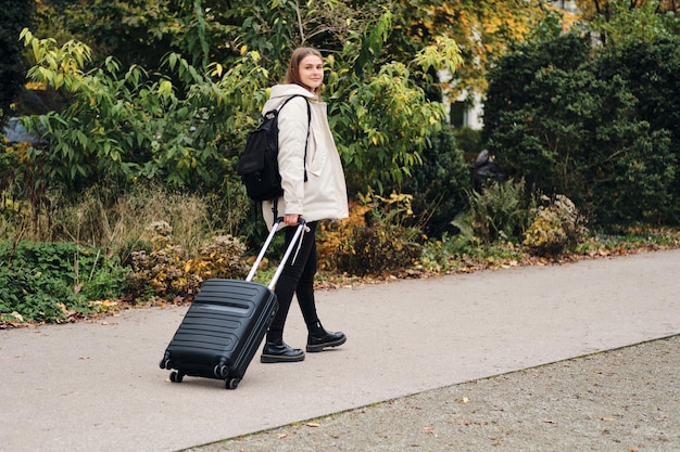 Hübsches lächelndes Mädchen in Daunenjacke, das mit Koffer und Rucksack glücklich durch den Stadtpark geht