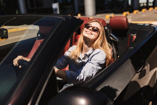 Hübsches lächelndes Mädchen mit Sonnenbrille, das glücklich in die Kamera schaut, während es Cabriolet-Auto auf den Straßen der Stadt im Freien fährt