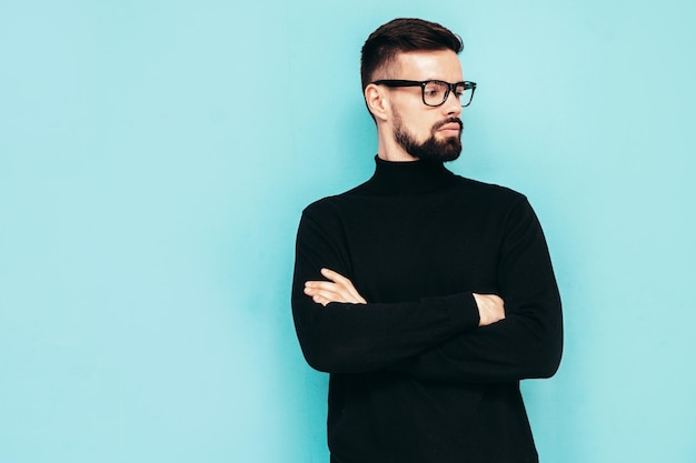 Hübsches lächelndes Modell Sexy stilvoller Mann in schwarzem Rollkragenpullover und Jeans gekleidet Mode-Hipster-Männchen posiert in der Nähe der blauen Wand im Studio Isoliert In Brille oder Brille Gekreuzte Arme