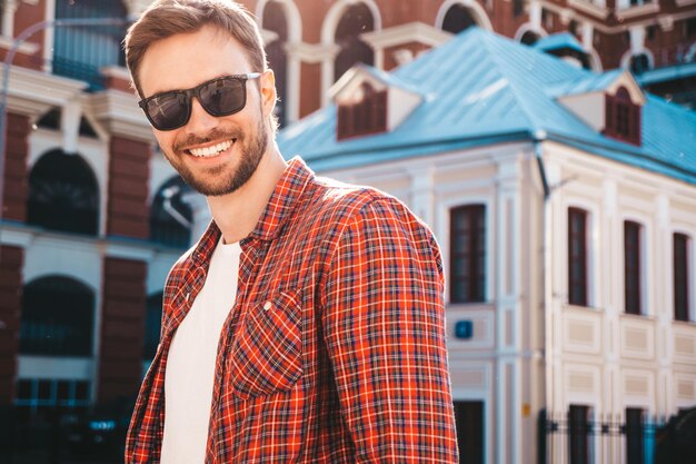 Hübsches lächelndes stilvolles Hipster-Lambersexual-Modell. Moderner Mann in rot kariertem Hemd. Modemann, der nahe Wolkenkratzer auf dem Straßenhintergrund in der Sonnenbrille aufwirft. Im Freien bei Sonnenuntergang