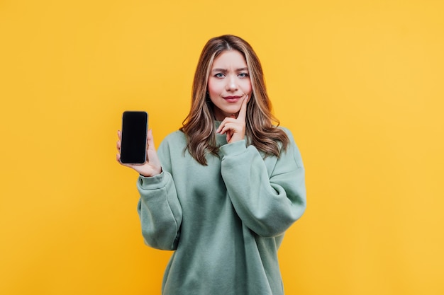 Hübsches Mädchen auf gelbem Hintergrund hält ein Telefon mit schwarzem Bildschirm und sieht nachdenklich aus