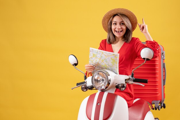 Hübsches Mädchen auf Moped mit rotem Koffer, der die Karte mit dem Finger nach oben hält