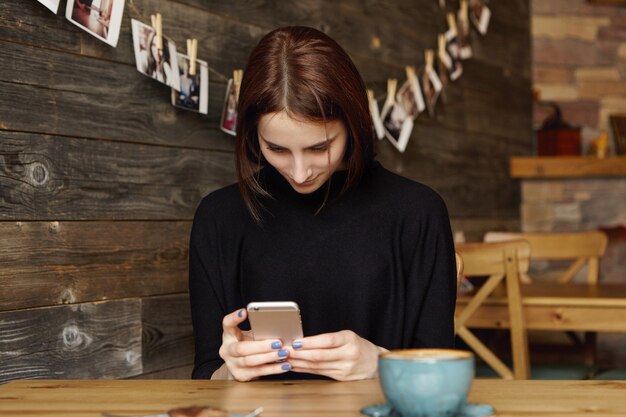 Hübsches Mädchen, das am Kaffeetisch mit Becher sitzt und drahtlose Internetverbindung auf Handy verwendet