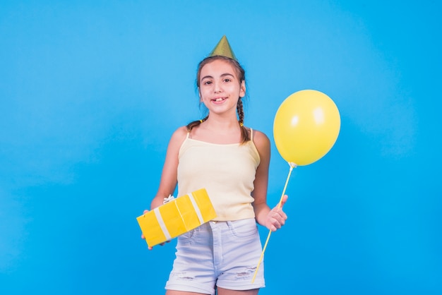 Hübsches Mädchen, das mit Geschenkbox und Ballonen auf blauer Tapete steht