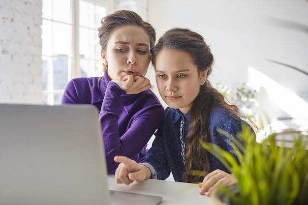 Hübsches Mädchen, das zu Hause vor offenem tragbarem Computer sitzt und Finger auf Bildschirm zeigt, Mutter neben ihr sich unsicher und schockiert fühlt