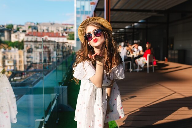 Hübsches Mädchen im Hut mit langen Haaren hört Musik durch Kopfhörer auf der Terrasse. Sie trägt ein weißes Kleid mit nackten Schultern, Sonnenbrille und rotem Lippenstift. Sie schickt einen Kuss an die Kamera.