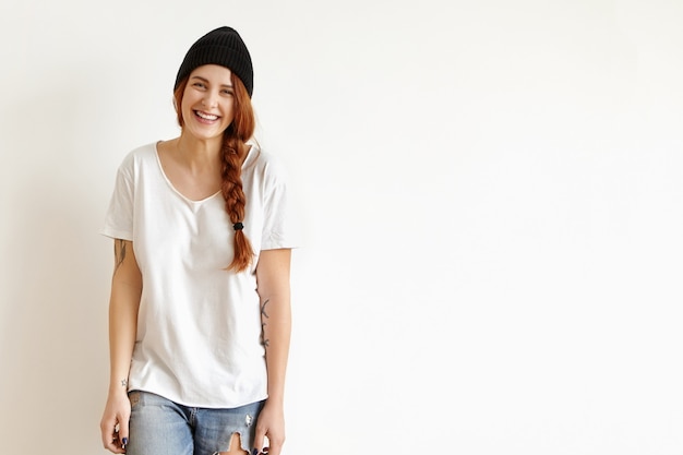 Hübsches Mädchen im stilvollen schwarzen Hut, im weißen T-Shirt und in den zerlumpten Jeans lächelnd, lokalisiert gegen Studiowand