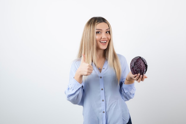 Hübsches Mädchen in blauem Outfit mit Purpurkohl, das Daumen aufgibt, auf Weiß.