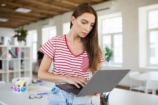 Hübsches Mädchen in gestreiftem T-Shirt, das nachdenklich einen Laptop mit modernem, leerem Büro im Hintergrund verwendet