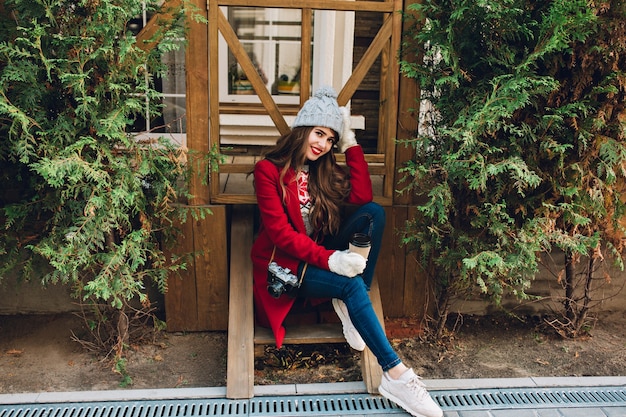 Hübsches Mädchen in voller Länge mit langen Haaren im roten Mantel und gestrickter Mütze, die auf Holztreppen im Freien sitzt. Sie hält Kamera und Kaffee in weißen Handschuhen und lächelt.