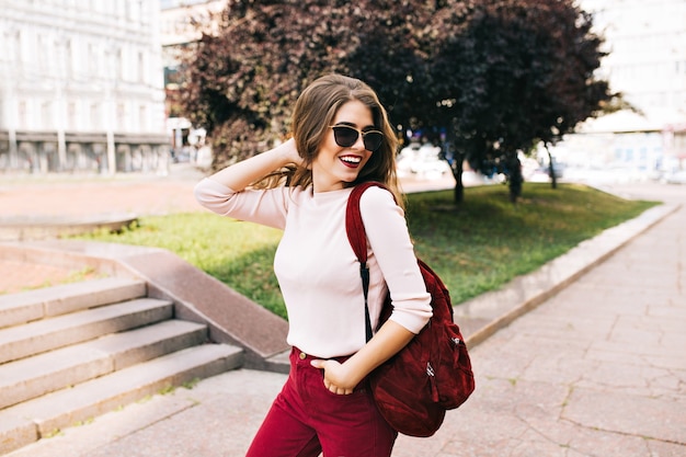 Hübsches Mädchen in weinigen Hosen in Sonnenbrille geht mit einer Tasche auf der Straße. Sie lächelt und sieht genossen aus.