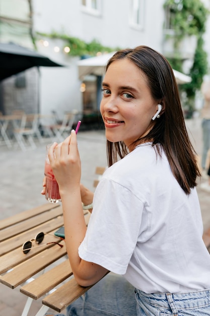 Hübsches Mädchen mit dunklem Haar in weißem T-Shirt mit Kopfhörern schaut in die Kamera und trinkt Smoothie, während es sich an warmen Tagen draußen in der Stadt ausruht