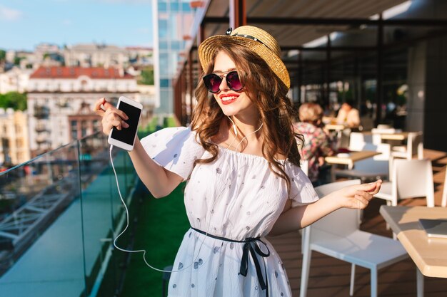 Hübsches Mädchen mit langen Haaren in der Sonnenbrille hört Musik durch Kopfhörer auf der Terrasse. Sie trägt ein weißes Kleid mit nackten Schultern, rotem Lippenstift und Hut. Sie tanzt.