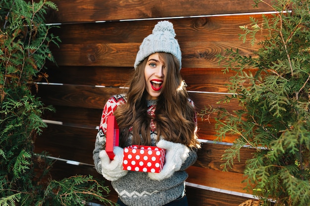 Hübsches Mädchen mit langen Haaren in der Winterkleidung auf Holz. Sie hält Weihnachtsgeschenk in Handschuhen und sieht erstaunt aus.