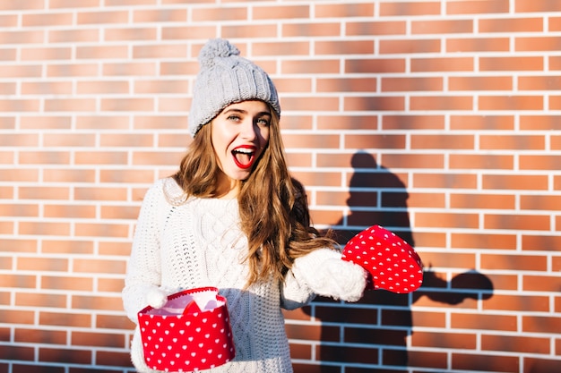 Hübsches Mädchen mit langen Haaren in Strickmütze und Handschuhen mit offenem Geschenk an der Wand draußen. Sie sieht erstaunt aus.