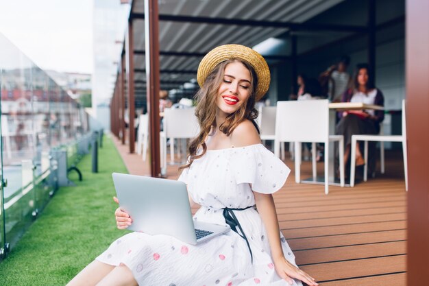 Hübsches Mädchen mit langen Haaren sitzt auf dem Boden auf der Terrasse. Sie trägt ein weißes Kleid mit nackten Schultern, rotem Lippenstift und Hut. Sie hält einen Laptop auf den Knien und lächelt.