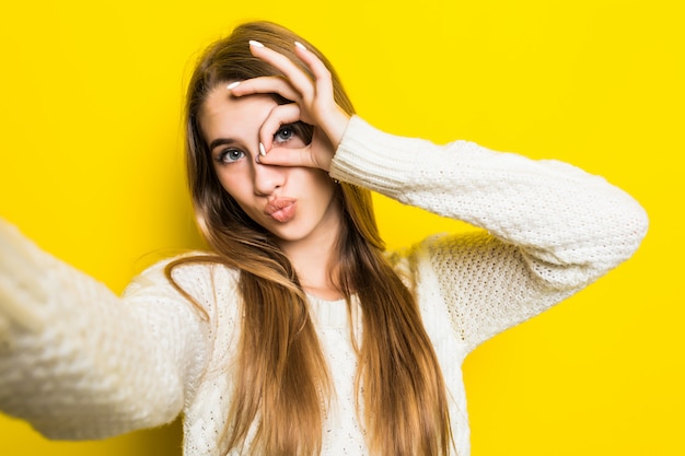 Hübsches modisches Mädchen macht Selfie auf ihrem Telefon und trägt einen breiten weißen Pullover auf Gelb