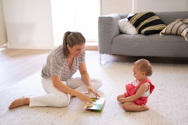 Hübsches Mutter-Lesebuch zum niedlichen kleinen Baby in roten Latzhose-Shorts. Konzentriertes Kleinkind, das auf Teppich im Wohnzimmer sitzt und Lesen lernt. Familie, Mutterschaft und Zuhause sein Konzept