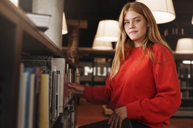 Hübsches rothaariges Mädchen, Student nimmt ein Buch aus dem Regal in der Bibliothek oder im Buchladen und lächelt in die Kamera.